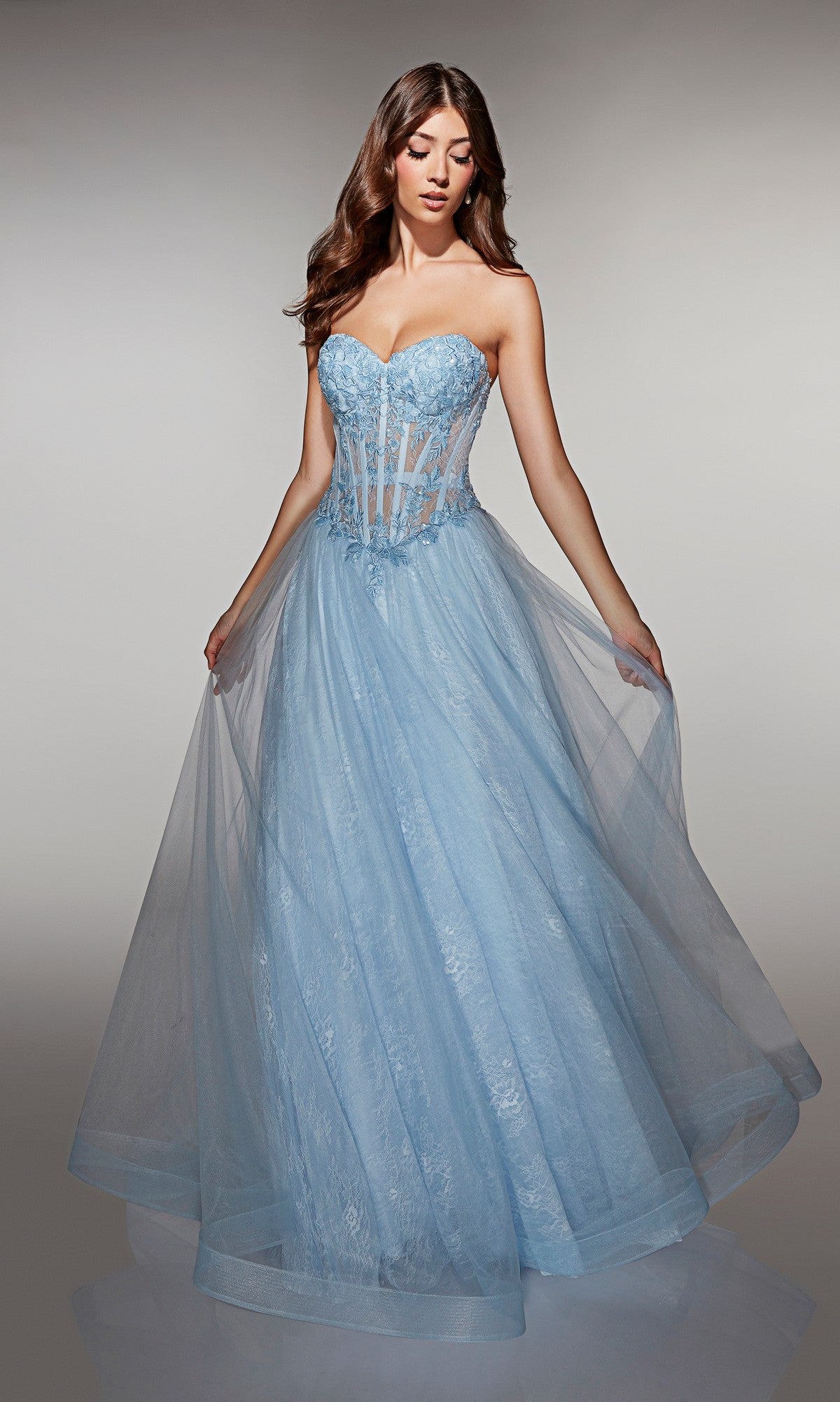 A woman in a glacier blue Alyce 61984 long prom dress with a strapless sheer corset bodice and full tulle ball gown skirt stands against a plain background.
